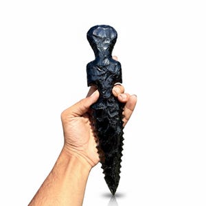 Könnte beinhalten: Ein schwarzer Obsidian-Dolch mit einem Griff in Form einer geschnitzten menschlichen Figur. Die Klinge des Dolches ist spitz und gezahnt. Der Obsidian hat eine glänzende, reflektierende Oberfläche. Der Dolch wird vor weißem Hintergrund gehalten.