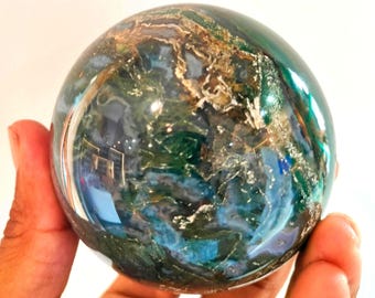 May include: A polished gemstone sphere with a mix of green, blue, and brown colours. The sphere has a reflective surface, and the colours swirl together in a natural pattern. The sphere is held in a hand.