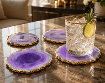 May include: A set of four circular purple agate coasters with irregular, textured gold-toned edges. One coaster supports a clear, faceted glass filled with ice, a lime slice, and a sprig of rosemary, showcasing the natural stone banding.
