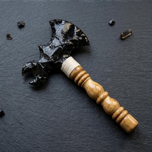 Può includere: Un'ascia in ossidiana fatta a mano con manico in legno. La testa in ossidiana nera è finemente scolpita e il manico è avvolto con spago bianco. Il manico in legno ha una finitura marrone chiaro. Piccoli frammenti di ossidiana sono sparsi.