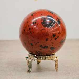 Può includere: Una sfera di ossidiana rossa lucida con inclusioni nere, esposta su un piccolo supporto in metallo dorato ornato. La sfera è un oggetto decorativo, probabilmente per l'esposizione o la collezione.