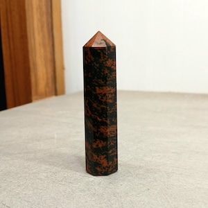 May include: A tall, hexagonal obsidian crystal point. The polished stone features a mix of black and reddish-brown colors. The crystal is standing upright on a light gray surface. The top of the crystal is a reddish-brown color.