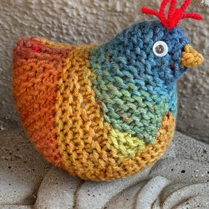 Op de afbeelding: Een gebreid regenboogkleurig kippen speelgoed met een rode kam en een wit knoop oog.
