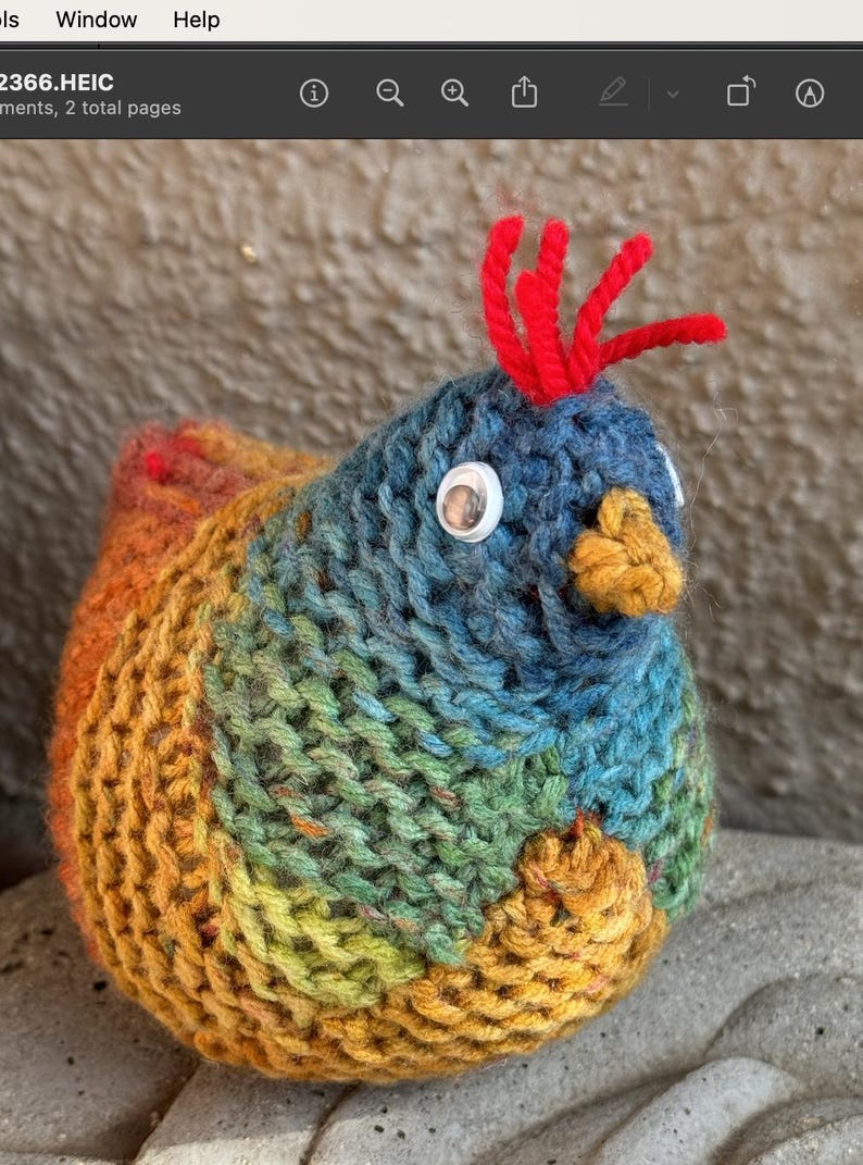 K&ouml;nnte beinhalten: Ein farbenfrohes gestricktes Vogelspielzeug mit roter Haube, wei&szlig;en Glitz Augen und einem gelben Schnabel. Der Vogel ist aus verschiedenen Garnfarben gefertigt, darunter Blau, Gr&uuml;n, Gelb und Orange.