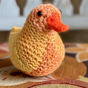May include: A handmade knitted duck toy in shades of yellow, orange, and peach. The duck has a bright orange beak and small, dark eyes. The knitted texture is visible, and the toy is sitting on a patterned surface.