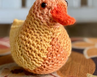 Peut inclure: Un jouet de canard tricoté à la main, aux tons de jaune, orange et pêche. Le canard a un bec orange vif et de petits yeux foncés. La texture tricotée est visible. Le jouet est posé sur une surface à motifs.