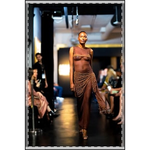May include: A model walks a runway wearing a sheer, brown, floor-length gown. The dress features a beaded bodice and a draped skirt with a high slit. The model is wearing gold heels. The background shows an audience.