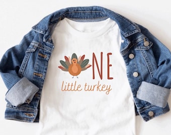 Può includere: Maglietta bianca con un'illustrazione di tacchino marrone e arancione e il testo "One little turkey".