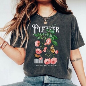 May include: Dark gray t-shirt with a floral design and the text "PATHOLOGICAL PEOPLE PLEASER". The design includes pink roses, green leaves, and butterflies. The shirt also has the text "DO SOMETHING RARE SAY SOMETHING".