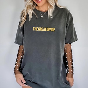 Pu&ograve; includere: T-shirt grigio scuro con la scritta "THE GREAT DIVIDE" in giallo. La maglietta &egrave; indossata con una maglia a maniche lunghe in rete nera con dettagli di perle bianche. La modella indossa una collana d'argento.