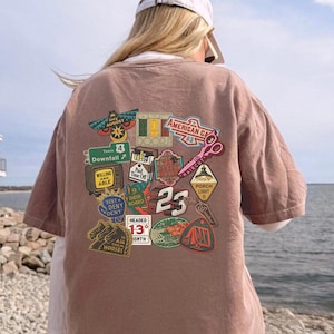 Pu&ograve; includere: Una maglietta marrone con una collezione di toppe grafiche colorate. Le toppe includono testi come "August", "Downfall", "American CA", "Haircut" e "23". La maglietta &egrave; indossata da una persona con un berretto da baseball bianco.