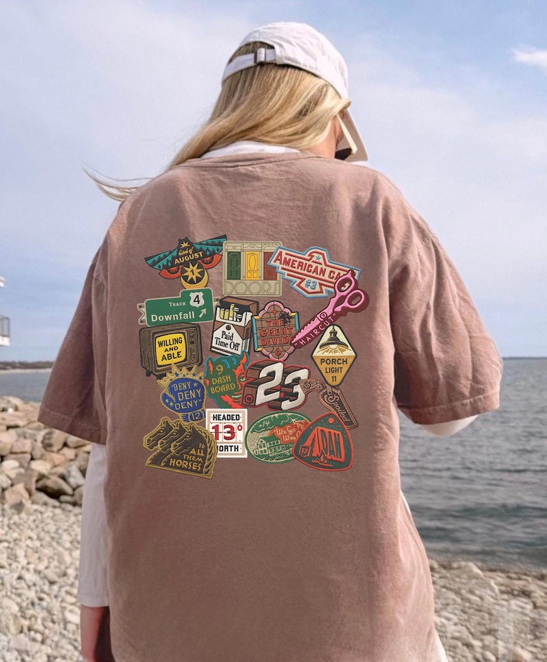 Pu&ograve; includere: Una maglietta marrone con una collezione di toppe grafiche colorate. Le toppe includono testi come "August", "Downfall", "American CA", "Haircut" e "23". La maglietta &egrave; indossata da una persona con un berretto da baseball bianco.