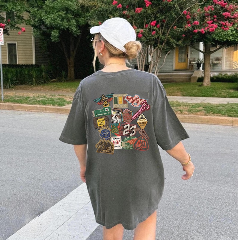 Pu&ograve; includere: T-shirt oversize grigio scuro con un collage di toppe colorate e il numero 23. Le toppe includono vari disegni, come segnali stradali, forbici e la scritta "American Car". La maglietta &egrave; indossata con un berretto da baseball bianco.
