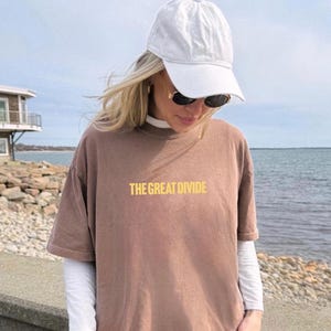 Pu&ograve; includere: Una maglietta marrone con la scritta "THE GREAT DIVIDE" in giallo. La maglietta &egrave; indossata sopra una maglia bianca a maniche lunghe. Sono visibili anche un berretto da baseball bianco e occhiali da sole.