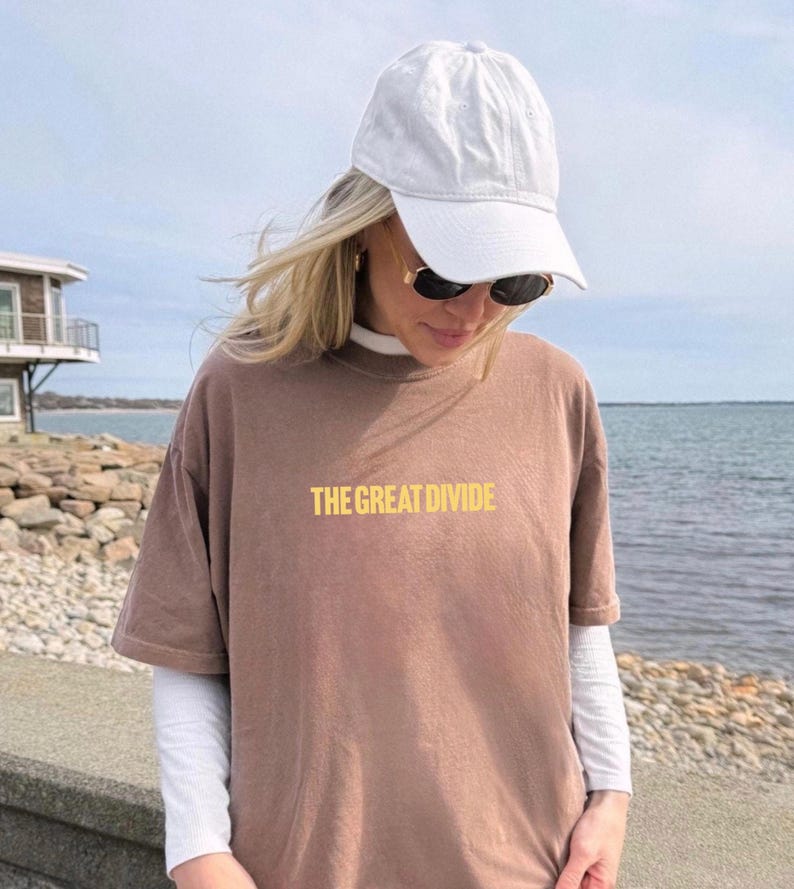 Pu&ograve; includere: Una maglietta marrone con la scritta "THE GREAT DIVIDE" in giallo. La maglietta &egrave; indossata sopra una maglia bianca a maniche lunghe. Sono visibili anche un berretto da baseball bianco e occhiali da sole.