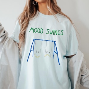 Puede incluir: Camiseta azul claro con la frase "MOOD SWINGS" en verde. Debajo, un gráfico de un columpio azul con dos asientos, uno con una cara sonriente y otro con una cara triste. Ropa casual.