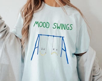 Può includere: T-shirt azzurra con la scritta verde "MOOD SWINGS". Sotto, un disegno di un'altalena blu con due seggiolini, uno con una faccia sorridente e l'altro con una faccia triste. Abbigliamento casual.