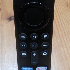 Puede incluir: Un mando a distancia negro con un pad de navegación circular y botones para varios servicios de streaming, incluyendo Prime Video, Netflix, DIRECTV y Peacock. El mando a distancia está en un soporte gris.