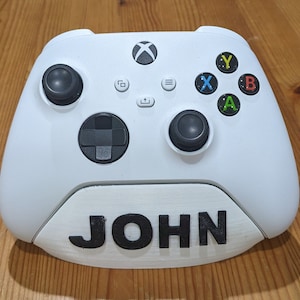 May include: White Xbox controller stand with the name "JOHN" printed on the front.
