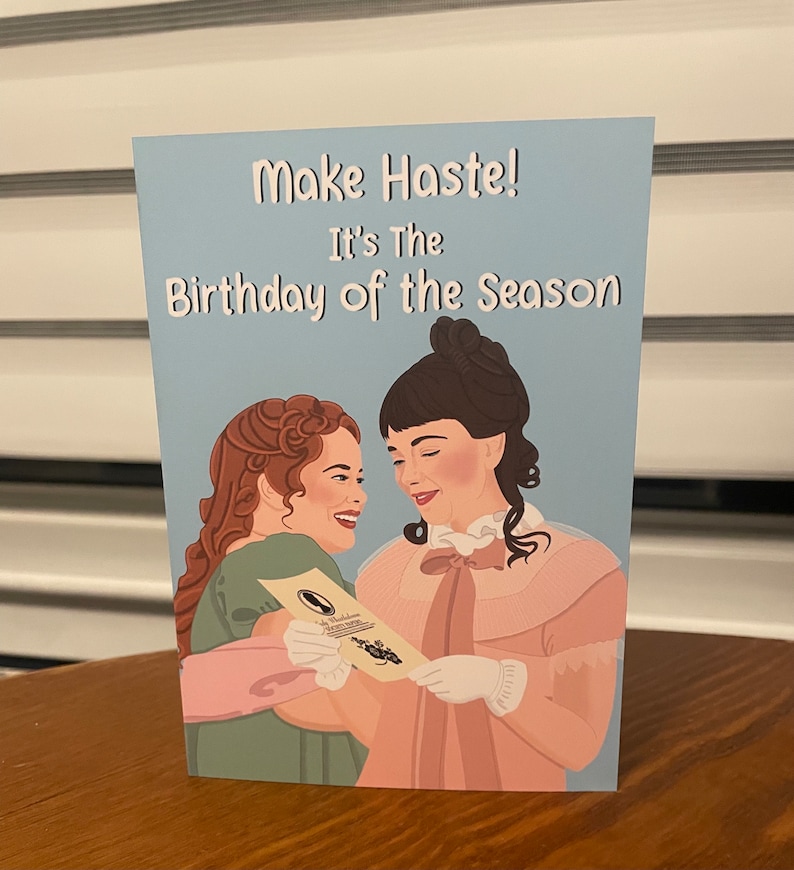 May include: A greeting card with a blue background featuring two women in period dress. The card reads "Make Haste! It's the Birthday of the Season."