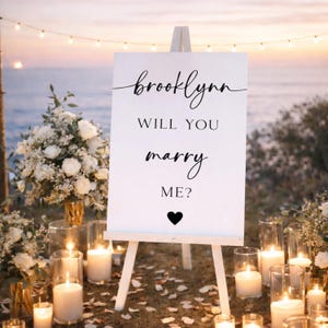 Puede incluir: Un letrero blanco sobre un caballete con las palabras "brooklynn, ¿te casarías conmigo?" en escritura negra. Un corazón negro en la parte inferior. La escena está decorada con velas, flores y luces de cadena, creando un ambiente romántico.