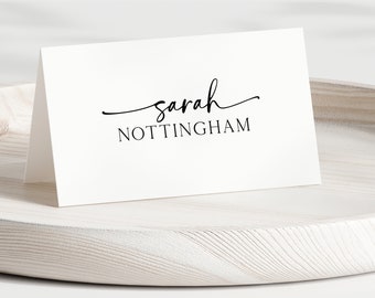 Può includere: Un segnaposto bianco piegato con il nome "Sarah" in corsivo sopra il cognome "NOTTINGHAM" in un carattere semplice. Il segnaposto è su un vassoio di legno chiaro.