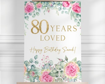 May include: A white sign on a wooden easel, decorated with pink roses and green foliage. The sign reads "80 YEARS LOVED" in gold lettering, with "Happy Birthday Sarah!" below. The sign is for a birthday celebration.