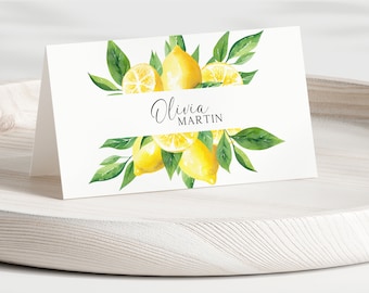 May include: A white folded place card featuring a watercolor illustration of yellow lemons and green leaves surrounding the printed black text Olivia MARTIN in a script and sans-serif font combination on the center white space.