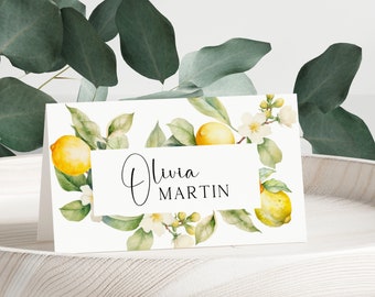 May include: A white folded place card featuring a watercolor illustration of yellow lemons, green leaves, and white blossoms. The center of the card displays the name Olivia Martin in a combination of script and sans-serif black lettering.