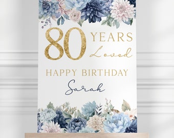 May include: A sign on an easel with a floral design and gold text. The sign reads "80 YEARS Loved HAPPY BIRTHDAY Sarah". The floral arrangement includes blue, white, and pink flowers.