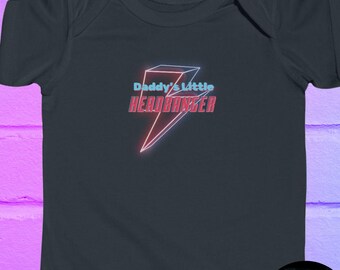May include: Dark gray baby bodysuit with the text "Daddy's Little Headbanger" in a neon-style design. The graphic features a lightning bolt shape with red and blue accents. A small, circular logo is visible at the bottom.