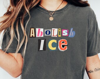 May include: Dark gray t-shirt with the text "Abolish ICE" in a colorful, cut-out letter design. The shirt is paired with blue jeans and white slip-on shoes. A casual, everyday look.