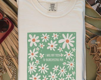May include: Cream-colored t-shirt with a green square featuring white and orange daisy graphics and the text "I WILL NOT YIELD TO DISRESPECTFUL MEN." The shirt is folded and has a Comfort Colors tag.