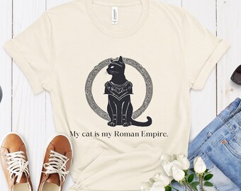 May include: A beige t-shirt with a black silhouette of a cat wearing a crown and sitting inside a circle. The text "My cat is my Roman Empire." is printed below the image.
