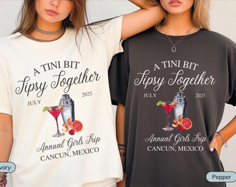 May include: Two white t-shirts with black text and graphics. The text on the shirts reads "A Tini Bit Sipsy Together" with the date "July 2025". The graphic is a cocktail shaker and a martini glass with a red drink. The text below the graphic reads "Annual Girls Trip Cancun, Mexico". One shirt is ivory and the other is pepper.