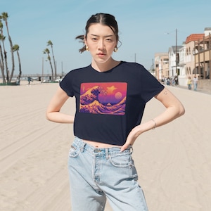 May include: A navy blue cropped t-shirt with a graphic print of the Great Wave off Kanagawa in a retro pixelated style. The wave is in shades of pink, purple, and yellow.