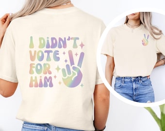 May include: Cream-colored t-shirt with the text "I DIDN'T VOTE FOR HIM" in pastel colors and a peace sign graphic. The peace sign and text have a rainbow gradient effect. The shirt is a casual style.