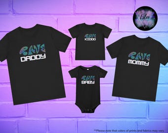 May include: Black t-shirts and a onesie with the words "RAVE DADDY", "RAVE KIDDO", "RAVE BABY", and "RAVE MOMMY" in white and a colorful design. The shirts are arranged on a purple and white brick wall.