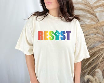 May include: Cream-colored t-shirt with the word "RESIST" in rainbow colors and a rainbow fist graphic. The shirt has short sleeves and a crew neck. The overall style is casual and comfortable.