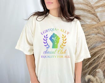 May include: Cream-colored t-shirt with a rainbow graphic that reads "LGBTQIA+ ALLY Social Club Equality For All." The design features a raised fist inside a laurel wreath. The shirt is a comfortable, oversized fit.