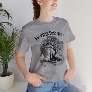 May include: A heather gray t-shirt featuring the text "Big River Conspiracy" above a black and white illustration of a weeping willow tree with figures beneath it. The shirt is a crew neck style.