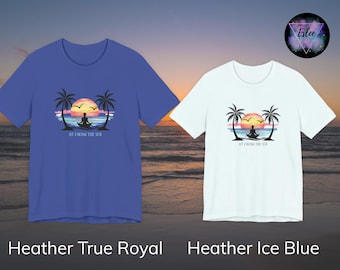 May include: Two t-shirts, one blue and one white, each featuring a sunset design with palm trees, a meditating figure, and the text "SIT FACING THE SEA." The blue shirt is labeled "Heather True Royal," and the white shirt is labeled "Heather Ice Blue."