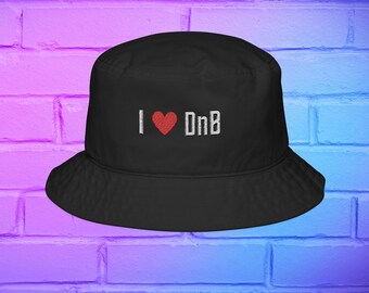 May include: Black bucket hat with the embroidered text "I ❤️ DnB" in white and red. The hat has a wide brim and a rounded crown. The hat is made of a soft material and is suitable for casual wear.