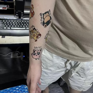 May include: A person's arm with several temporary tattoos of Pokemon characters. The tattoos include a pink Jigglypuff, a yellow Eevee, and a blue Snorlax. The tattoos are on the person's forearm and upper arm. The tattoos are in Japanese.