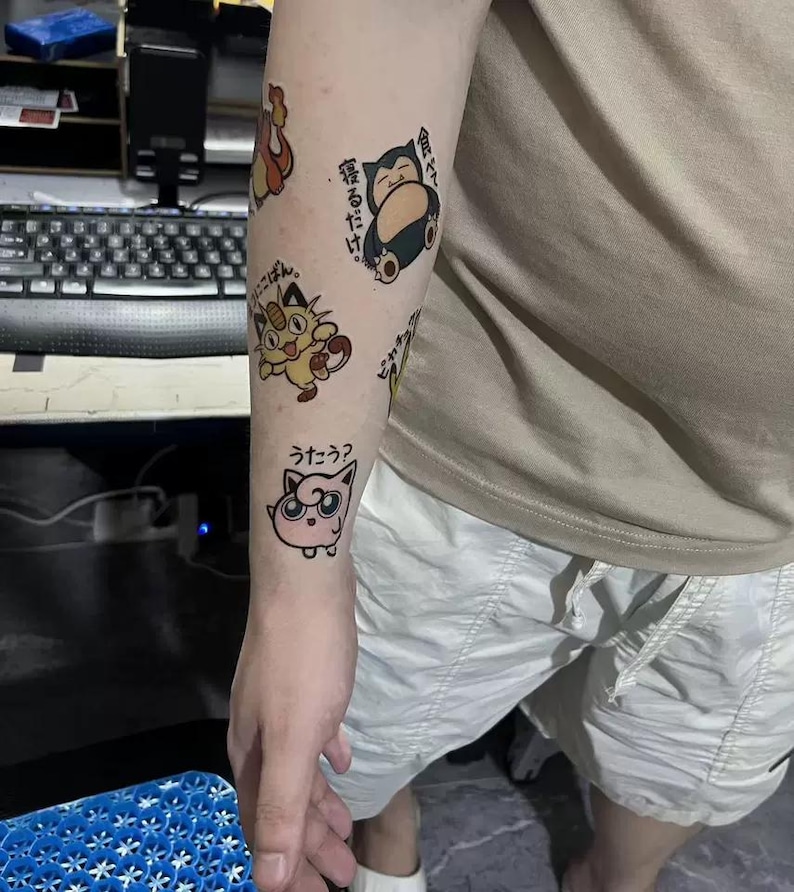 May include: A person's arm with several temporary tattoos of Pokemon characters. The tattoos include a pink Jigglypuff, a yellow Eevee, and a blue Snorlax. The tattoos are on the person's forearm and upper arm. The tattoos are in Japanese.