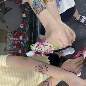 May include: Two children's arms with temporary tattoos of Pokemon characters. The tattoos feature Squirtle, Pikachu, and Jigglypuff. The tattoos are in Japanese and English.