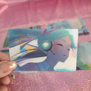 Puede incluir: Una tarjeta holográfica con una ilustración de estilo anime de una mujer con cabello azul usando auriculares. El fondo es una imagen borrosa de una ciudad.