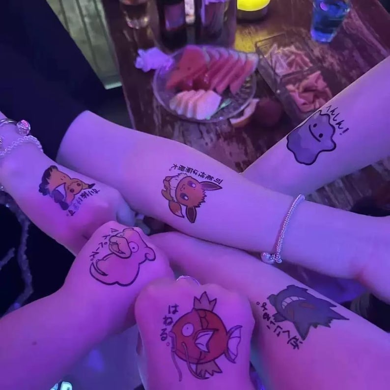 May include: Close-up of four arms with temporary tattoos of cartoon characters from the Pokemon franchise. The tattoos are in Japanese and feature characters like Pikachu, Squirtle, and Gyarados. The tattoos are on a light skin tone.