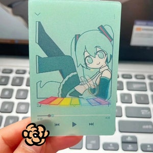 May include: A translucent green card with a cartoon illustration of a female anime character with long green hair. The character is sitting on a rainbow-colored platform. The card has a music player interface with the time 0:39 and 4:15 displayed.