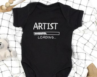 May include: Black baby onesie with the text "ARTIST LOADING..." in white. The onesie is displayed on a white and black checkered blanket, with a small teddy bear and wooden beads in the background.